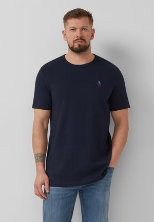 Man with short hair and beard wearing a navy blue t-shirt with a small embroidered skier and light blue jeans, standing against a gray background.