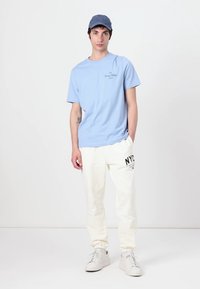 T-shirt bleu clair à manches courtes avec un petit logo sur la poitrine, associé à un pantalon de jogging crème avec un imprimé NYC, et des baskets blanches.