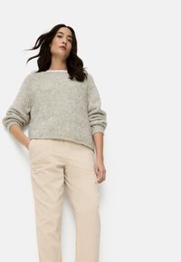 Femme portant un pull en maille gris clair et un pantalon beige, debout avec une main dans la poche, devant un fond blanc uni.
