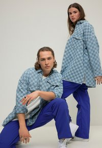 Due modelli posano in camicie di denim a scacchi blu e pantaloni viola, mostrando trame e motivi contrastanti su uno sfondo neutro.