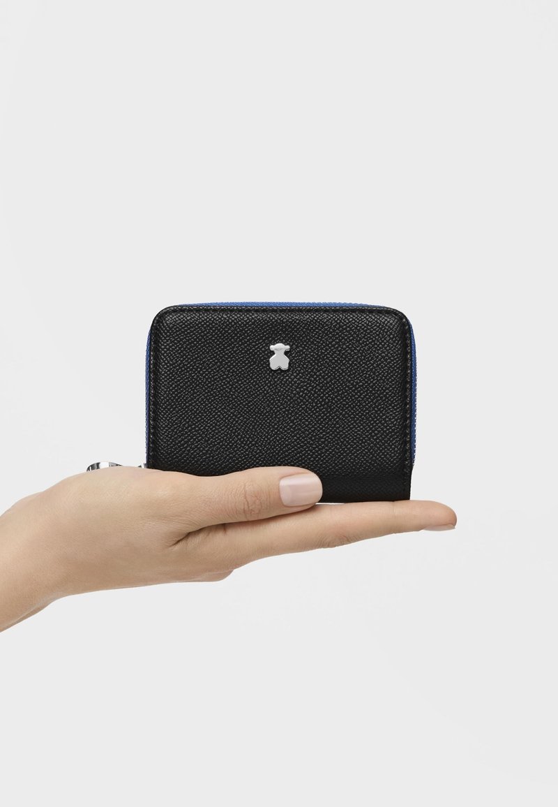 Portafoglio nero con texture, con dettagli in blu sulla cerniera e logo di un orso argentato, tenuto in mano. Forma rettangolare compatta per una facile maneggevolezza.
