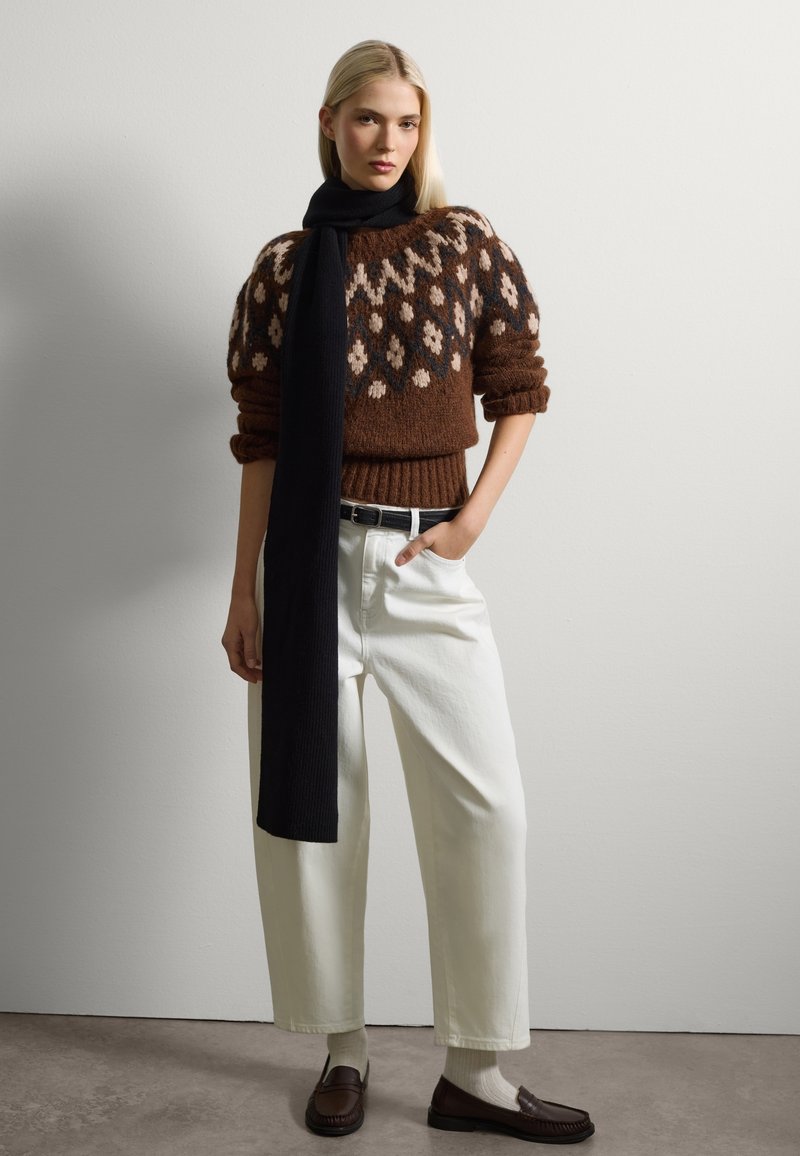 Brauner gemusterter Pullover mit Creme- und schwarzen Akzenten, weiße hochgeschnittene Hose, schwarzer Schal und braune Slipper. Glatte Textur, lockere Passform.