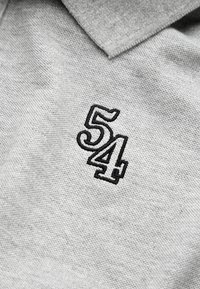 Grå polo shirt med et tekstureret stof, prydet med et sort broderet '54' på venstre side. Klassisk kravedesign.