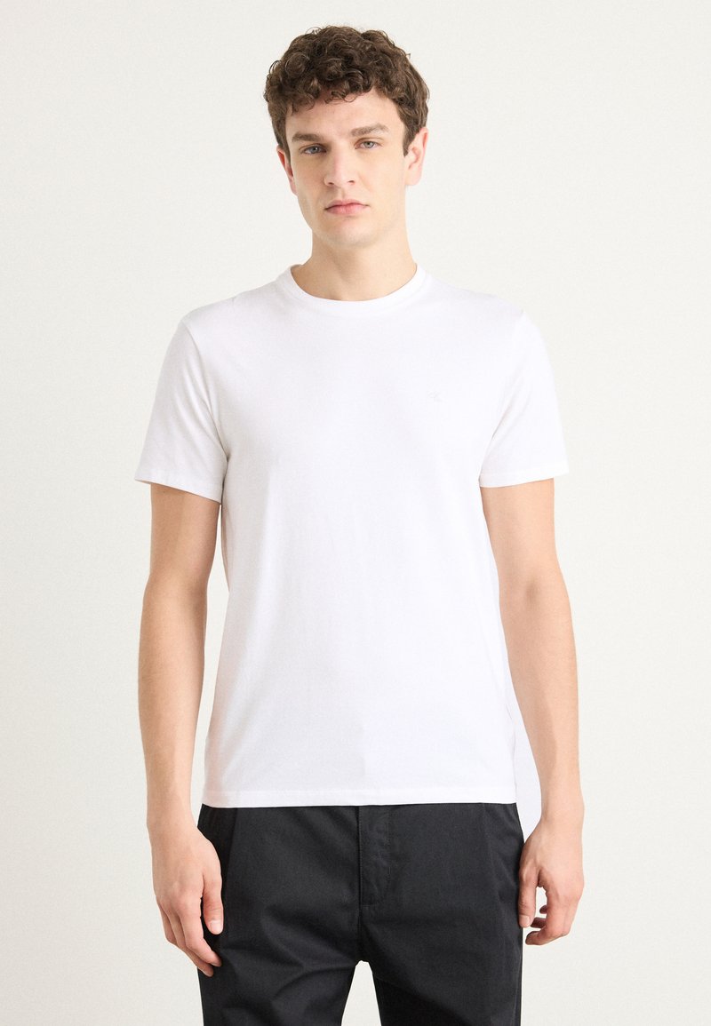 Witte katoenen t-shirt met korte mouwen en een ronde hals, gladde textuur, met een subtiel logo op de borst. Gecombineerd met donkere broeken.