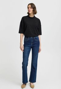 Schwarzes, strukturiertes Kurzarmoberteil mit hohem Kragen, kombiniert mit dunkelblauen Schlagjeans und beigen Flachschuhen. Schlichtes, modernes Design.