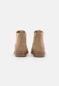 Bottines en suède marron avec des panneaux latéraux élastiques, surface texturée et un talon empilé. La vue arrière montre les détails de couture et l'onglet.