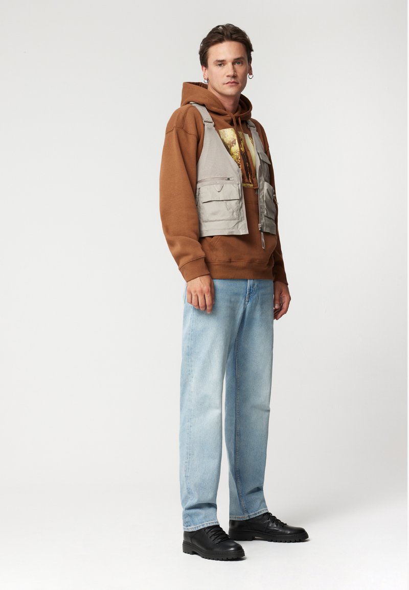 Jasnoszara kamizelka robocza noszona na brązowej bluzie z kapturem, z kieszeniami. Uzupełniona jasnoniebieskimi dżinsami i czarnymi sznurowanymi butami. Prosty design.