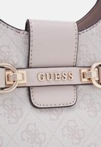 Beige mønstret håndtaske med en glat læderrem og guldfarvet hardware. Har "GUESS" logoet på en dekorativ flap.