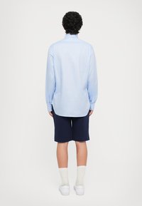 Světle modrá košile s dlouhými rukávy, doplněná námořnickými šortkami. K outfitu patří bílé ponožky a tenisky. Zobrazeno zezadu.