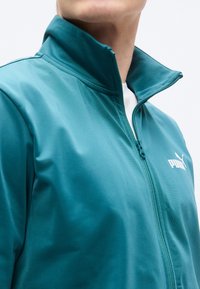 Μπουφάν σε πετρόλ χρώμα με φερμουάρ και ψηλό γιακά, καθώς και πλαϊνές τσέπες· κατασκευασμένο από λεία, ελαφριά ύφασμα· διαθέτει λευκό λογότυπο στο στήθος.