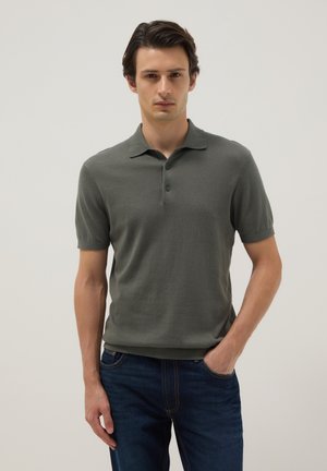 Hombre con una camisa polo de manga corta verde oliva y jeans azul oscuro, de pie con una mano en el bolsillo frente a un fondo liso.