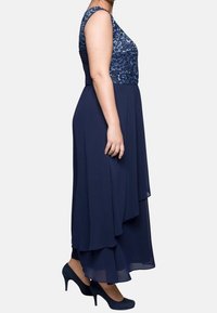 Robe bleu marine avec un corsage ajusté à motif floral et une jupe fluide et superposée. Portée avec des talons assortis bleus. Modèle sans manches.