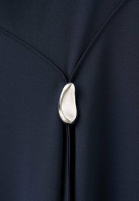 Nawojowy detal materiału z gładkim, polerowanym srebrnym akcentem dekoracyjnym w kształcie kropli, podkreślającym design odzieży.