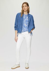 Blauwe bomberjack, blauw-wit gestreepte top, strakke witte jeans en beige sneakers. De jas heeft een rits, elastische mouwen en een verstelbare zoom.