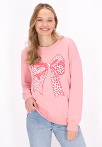 Felpa rosa con una grande grafica a fiocco in stampa leopardata, maniche lunghe e vestibilità comoda. Indossata con jeans azzurri.