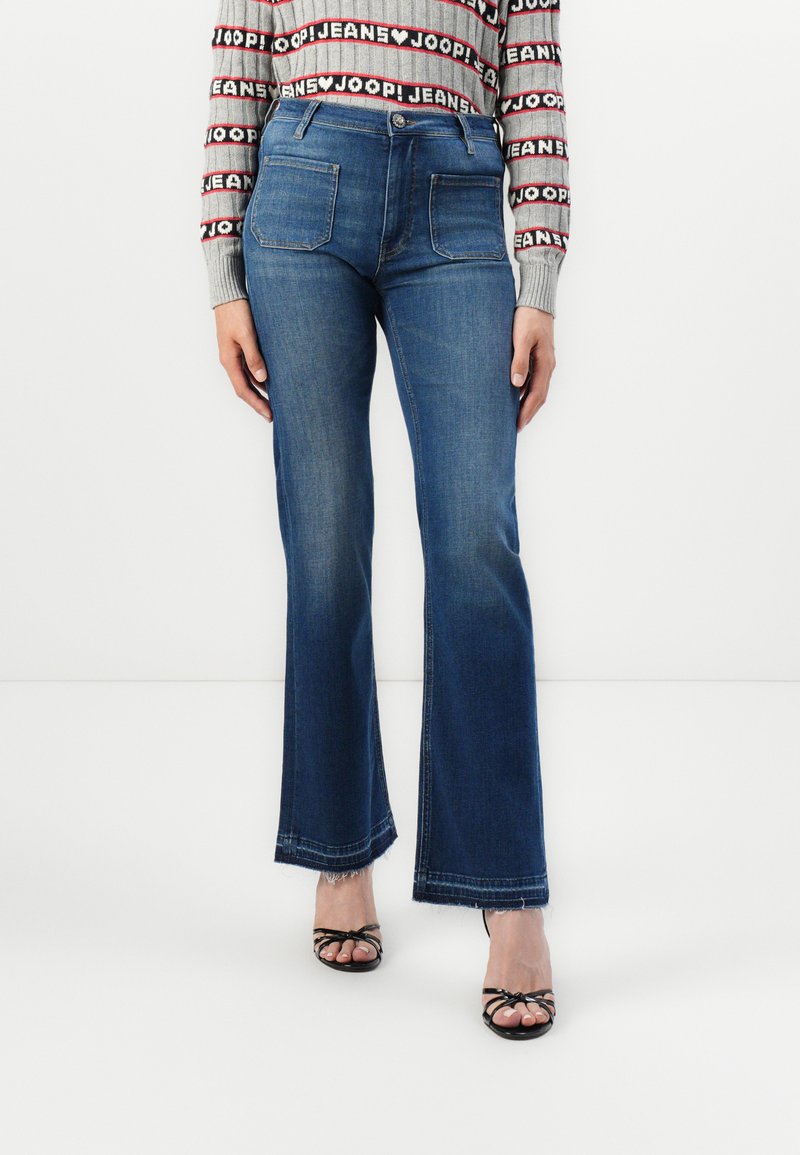 Dunkelblaue, ausgestellte Jeans mit mittlerer Leibhöhe, fünf Taschen und kontrastierenden Nähten. Kombiniert mit schwarzen Riemchensandalen.