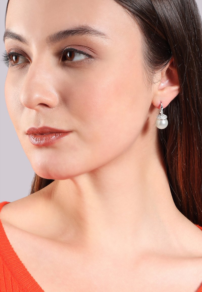 Silberne Ohrringe mit einem runden Perlenanhänger und einem Kristallakzent, vor einem neutralen Hintergrund präsentiert. Die Trägerin hat glattes Haar.