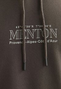Braune Kapuzenjacke mit einem Frontdruck, auf dem steht "MENTON 43° 42' 30'N 7° 30' 00'E Provence-Alpes-Côte d'Azur" und geflochtenen Kordeln mit Kunststoffspitzen.