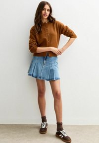 Kvinna iklädd en brun knäppt cardigan, ljusblå denimfållad kjol, bruna strumpor och bruna sneakers med vita snören, stående mot en vit vägg.