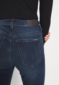 Persona con jeans de Calvin Klein de color azul oscuro y un suéter negro de canalé, con la mano en la cadera, vista desde atrás.