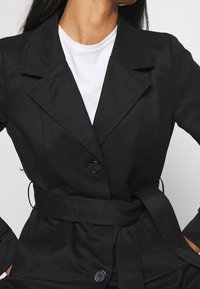 Mujer vestida con un abrigo negro de trench con cinturón sobre una camiseta blanca de cuello redondo, con las manos en los bolsillos del abrigo, frente a un fondo liso.