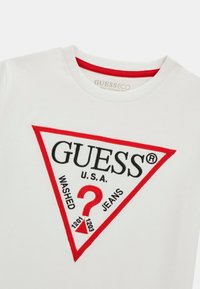 Balts kokvilnas T-krekls ar apaļu kaklu, ar izteiksmīgu sarkanu un melnu trīsstūra logo, uz kura ir uzraksti "GUESS," "?" un "WASHED JEANS".