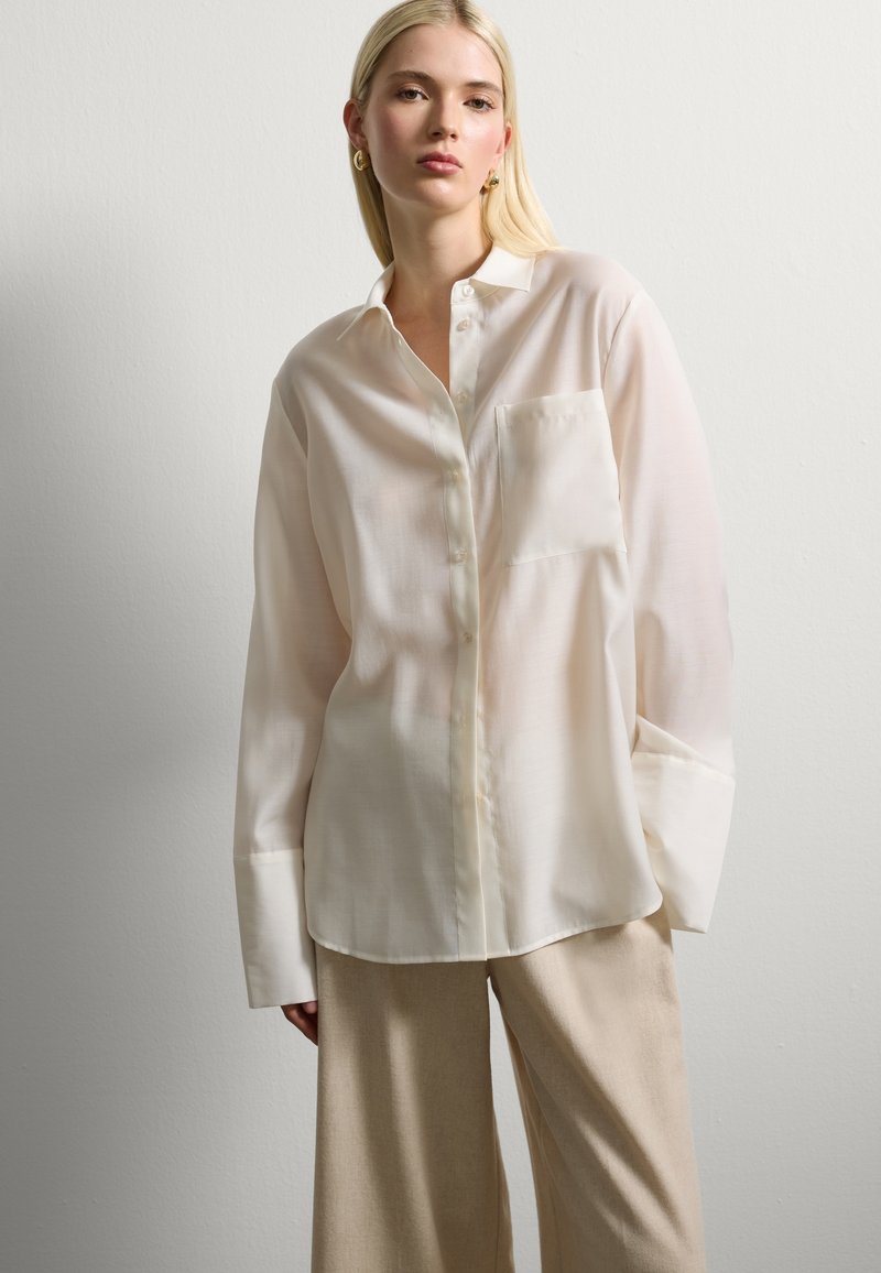 Blusa a maniche lunghe beige chiaro realizzata in tessuto liscio, con una chiusura frontale a bottoni, maniche con polsini e una tasca sul petto.