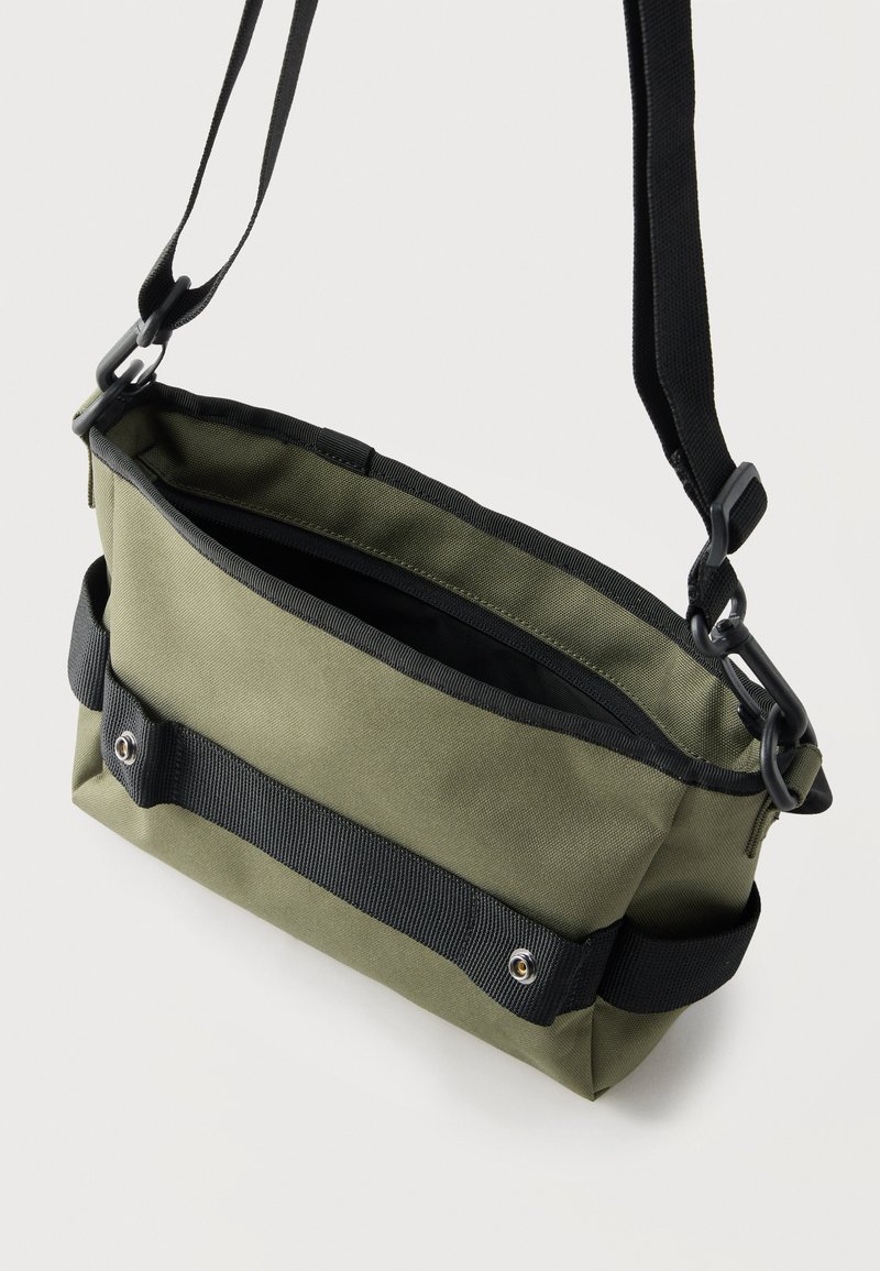 Groene canvas schoudertas met zwarte verstelbare riem, vooraan een horizontale zwarte riem met drukknopen en ritssluiting.
