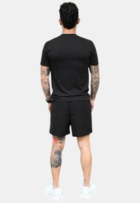 Camisa negra de manga corta y pantalones cortos negros. La persona tiene tatuajes visibles en una pierna y lleva zapatillas deportivas blancas.