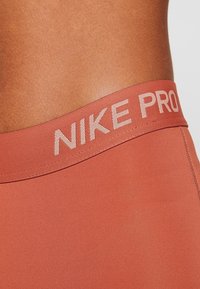 Legíny Nike Pro mají hladký, elastický materiál v barvě terakoty s kontrastním logem v světle béžové barvě na pasu.