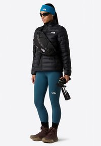 Czarna puchowa kurtka, turkusowe legginsy, brązowe buty trekkingowe oraz czarna saszetka na biodra. Niebieska opaska na głowę i okulary przeciwsłoneczne dopełniają outdoorowy look.