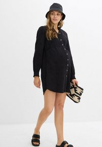 Donna incinta che indossa un abito nero con bottoni e un cappello a secchiello, che tiene una pochette a fantasia bianco e nero, mentre cammina con sandali neri.