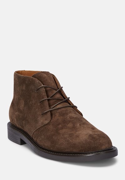 Botas de tobillo de ante marrón con punta redonda, cierre de cordones y suela plana oscura. Presentan una textura suave y un detalle de costura mínimo.
