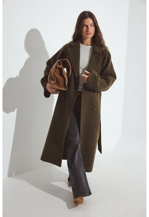 Oliivroheline villasegu mantel, millel on üleriietusele iseloomulik üleliigne lõige, kaks nuppu, koos halli särgi, tumesiniste laia säärega pükste ja pruuni nahast käekotiga.