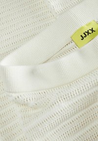 Bílý pletený svetr s texturovaným otevřeným tkaním, žebrovanými manžetami a viditelnou žlutou nášivkou s nápisem "JJXX".