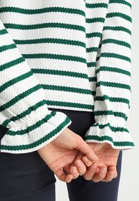 Kädet neulotut selän takana, päällä valkoinen, tekstuuriltaan epätasainen neulepaita, jossa on vihreät vaakaraitat, ja tummat housut.