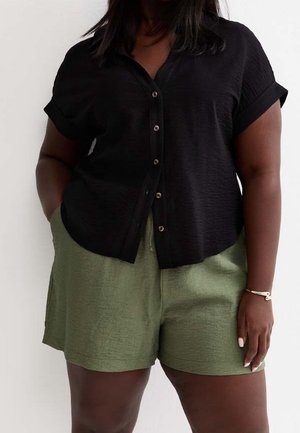 Femme portant une chemise noire à manches courtes boutonnée et un short vert taille haute, mains dans les poches sur fond blanc.