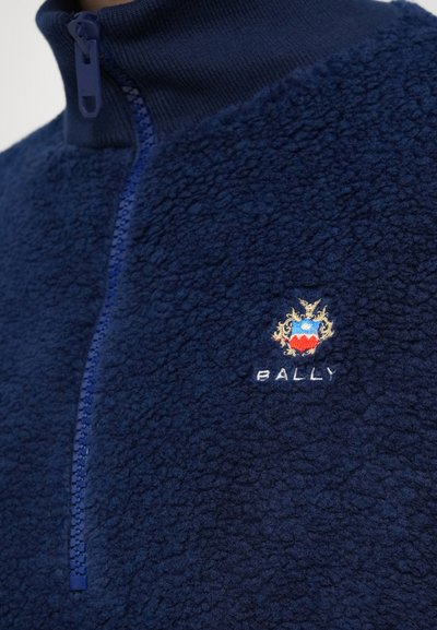 Tumši zilās vilnas mikas ar augstu apkakli un rāvējslēdzēja aizdari, ar strukturētu virsmu un izšūtu Bally logotipu zelta un sarkanā krāsā.