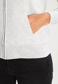 Sudadera con capucha gris con cierre de cremallera, confeccionada en una tela suave y texturizada, con dos bolsillos laterales y una cintura ajustada, visible sobre unos vaqueros oscuros.