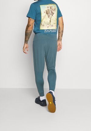 Turkusowe spodnie sportowe o dopasowanym kroju i teksturowanym pasie, zestawione z niebieskim t-shirtem z nadrukiem przedstawiającym roślinę pustynną i tekst.