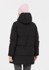 Sort puffersjakke med hætte, der har et kviltet design og elastiske manchetter. Bæret over grå leggings med en lyserød strikket hue.