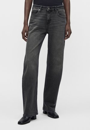 Personne portant un jean noir délavé à coupe droite et des bottines noires à bout carré, debout devant un fond clair uni.
