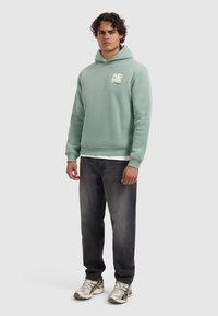 Man staat met een lichtgroene hoodie met grafische tekst, donker gewassen jeans en witte hardloopschoenen op een effen achtergrond.