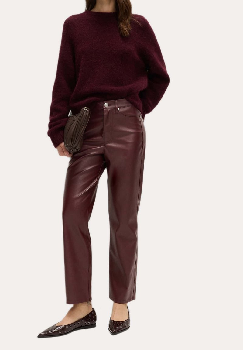 Femme portant un pull en peluche bordeaux, un pantalon en cuir bordeaux taille haute, tenant une pochette marron, et portant des ballerines noires pointues avec des œillets.