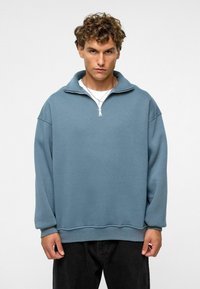 Lichtblauwe pullover sweatshirt met een ritssluiting op de kraag, ribbelstructuur en een ontspannen pasvorm. Gecombineerd met zwarte broeken voor contrast.