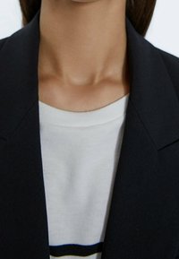 Černá sako s ostrými klopami nošené přes bílé triko s kulatým výstřihem, které má horizontální pruhy v námořnické modré a černé. Pohled na výstřih.