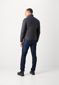 Veste matelassée gris foncé avec des coutures horizontales, un col haut, portée avec un jean bleu et des chaussures noires. Le tissu semble lisse et léger.