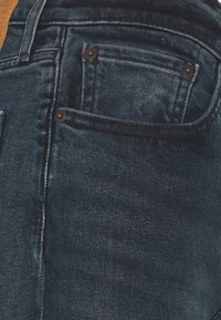 Jeans de mezclilla azul oscuro con diseño de cinco bolsillos, costuras en contraste y accesorios en tono cobre. Textura suave con un ligero lavado vintage.