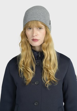 Blonde vrouw met lang golvend haar, die een grijze gebreide muts en een marineblauwe knopenjas draagt tegen een effen achtergrond.