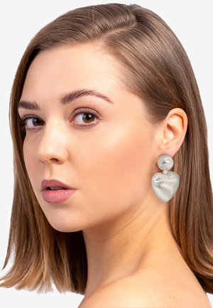 Silberne ohrringe in Herzform mit glänzender Oberfläche, versehen mit einem runden Oberaccent; vor einem neutralen Hintergrund präsentiert.